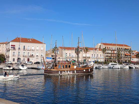Bateau de style ancien de l'association "Pirates" (c'est écrit sur la coque) vogue sur un canal à Sète.
Il y a des figures de pirates sur le pont et un cadeau de sauvetage moderne est attaché au navire.
Le ciel est très bleu, les façades claires un peu éblouissante