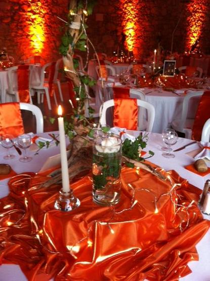 Salle de fête avec des tables brillamment décorées. Les murs en pierres sont éclairés en orange. Il y a du tissu orange qui brille en centre table pour supporter la décoration florale, tissu mis sur les chaises également et en ronds de serviettes