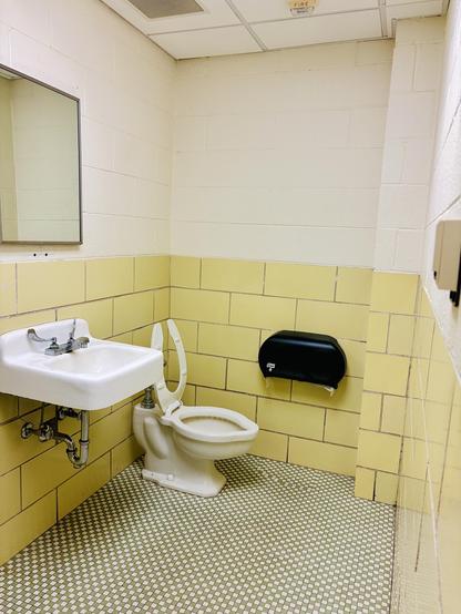 The image shows a small, utilitarian bathroom with yellow walls and a white floor. There is a white sink with a metal faucet and a white toilet with a black tissue dispenser mounted on the wall. The bathroom appears to be in a public facility, possibly a school or office building.