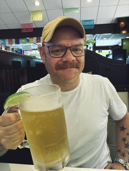 A man wearing a white t-shirt and a beige baseball cap is holding a large glass filled with a light-colored beverage, possibly beer, with a lime wedge on the rim. The background features a bar or restaurant setting with various decorations and a ceiling with colorful paper flags.