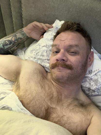 A man with a tattoo on his arm is lying in bed, wearing a white shirt with a floral pattern. He has a mustache and is bare-chested. The bed has a gray headboard and a white blanket.