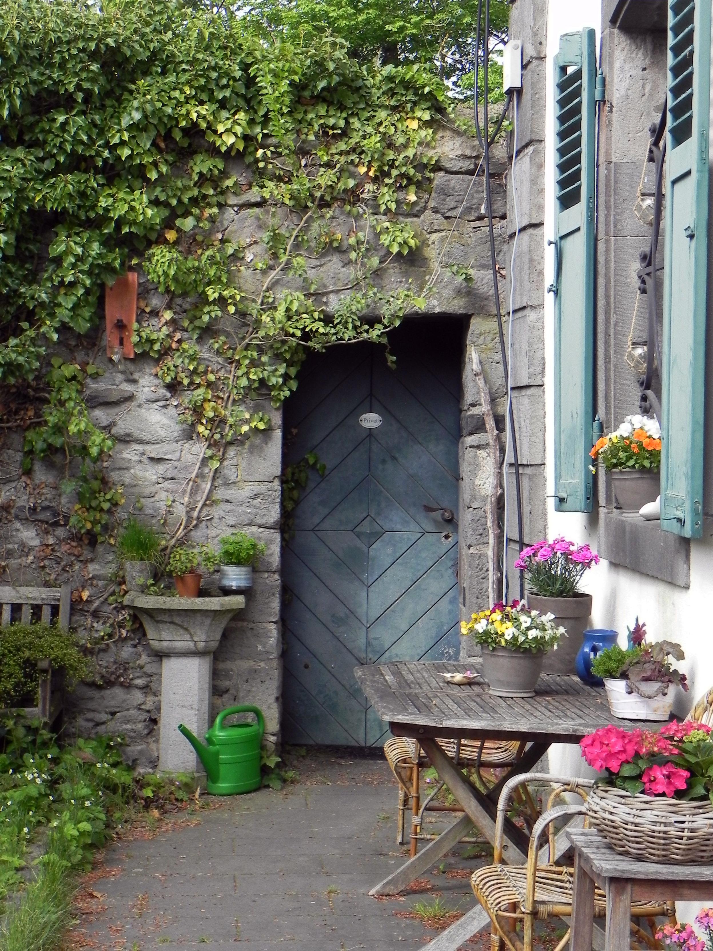 Une porte bleue en bois au fond d'une petite cour bien fleurie et verdoyante. Des pots et corbeilles de fleurs sont posés sur des tables et appui de fenêtre. Des volets en bois peints en bleu sont ouverts. Sur le mur, une plante verte grimpante le couvre en partie jusque tout près de la porte. Un nichoir est également visible. Au sol, il y a un arrosoir en plastique vert à côté d'un ancien bac à eau en pierre.

A blue wooden door at the end of a small, flowery and green courtyard. Pots and baskets of flowers are placed on tables and window sills. Wooden shutters painted blue are open. On the wall, a climbing green plant partly covers it up to very close to the door. A nest box is also visible. On the ground there is a green plastic watering can next to an old stone water tray.