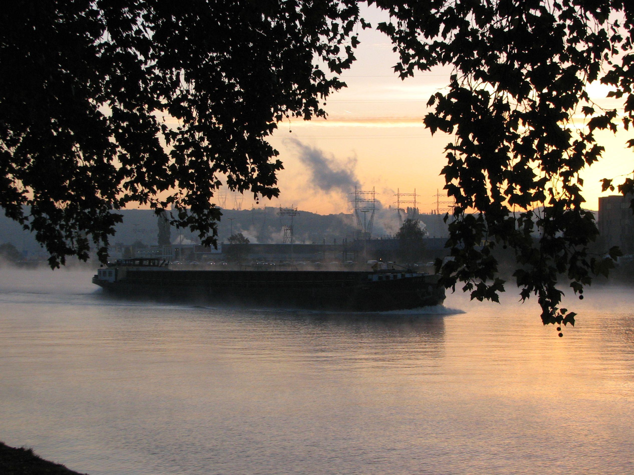 Une péniche sur La Meuse au point du jour, des fumerolles s'élèvent du fleuve du fait de la fraîcheur du matin. Un arbre cache partiellement la vue avec son feuillage. A l'arrière-plan, le ciel rougit à l'endroit où va se lever le soleil. On peut voir des collines boisées et de grands pylônes à haute-tension. De la fumée s'échappe d'une usine sur l'autre rive. 

A barge on the Meuse at daybreak, fumaroles rise from the river due to the coolness of the morning. A tree partially hides the view with its foliage. In the background, the sky turns red where the sun will rise. Wooded hills and large high-voltage pylons can be seen. Smoke escapes from a factory on the other bank.