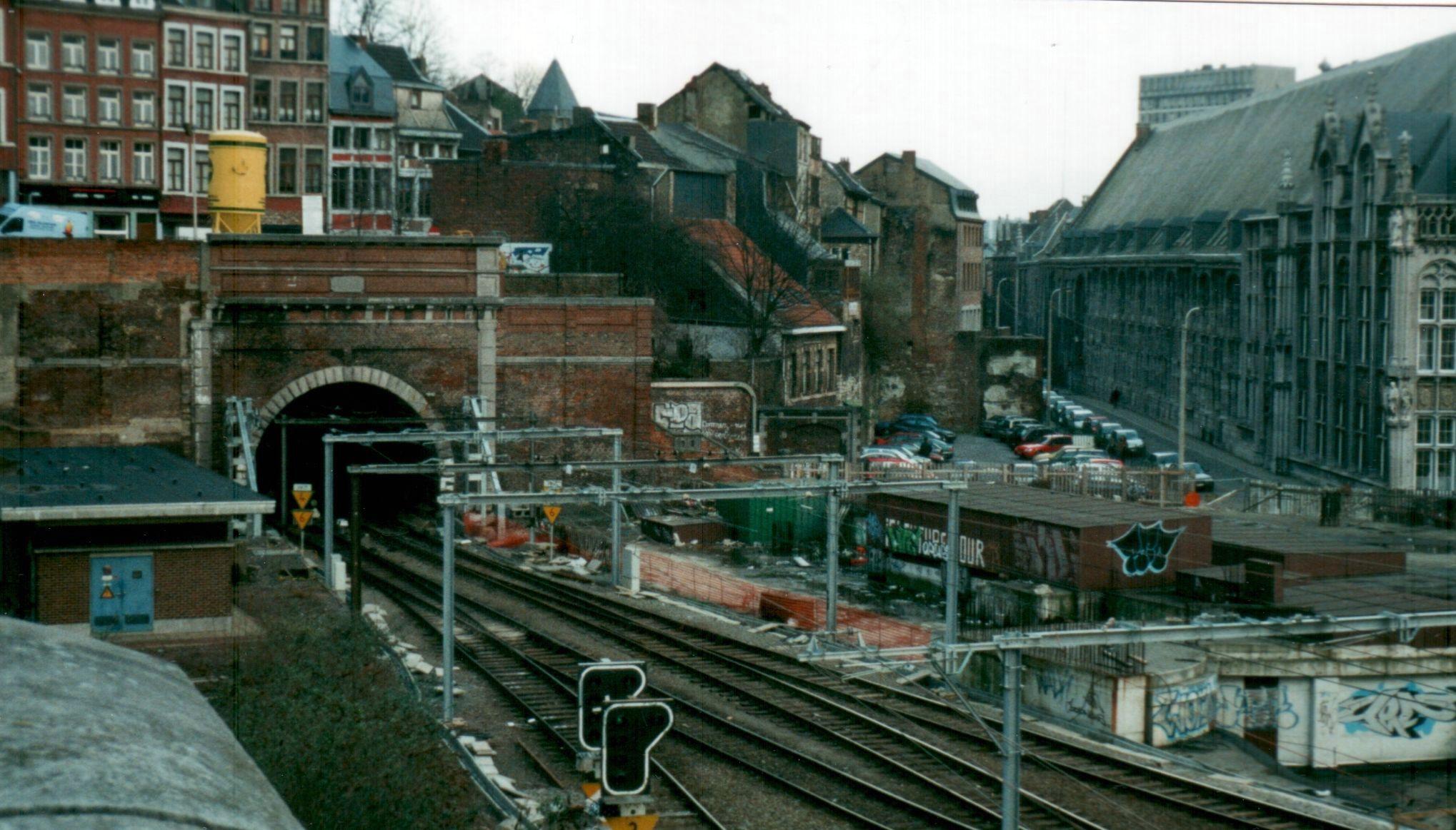 Des voies de chemin de fer qui entrent dans un tunnel dans un environnement urbain en chantier. A droite, on peut voir une partie du Palais des Princes Évêques (devenu palais provincial et de justice) auquel la gare devait son nom. Au-dessus du tunnel, des maisons anciennes en escalier. 

Railway tracks that enter a tunnel in an urban environment under construction. On the right, we can see part of the Palace of the Princes Bishops (now the provincial and courthouse) to which the station owed its name. Above the tunnel, old stepped houses.