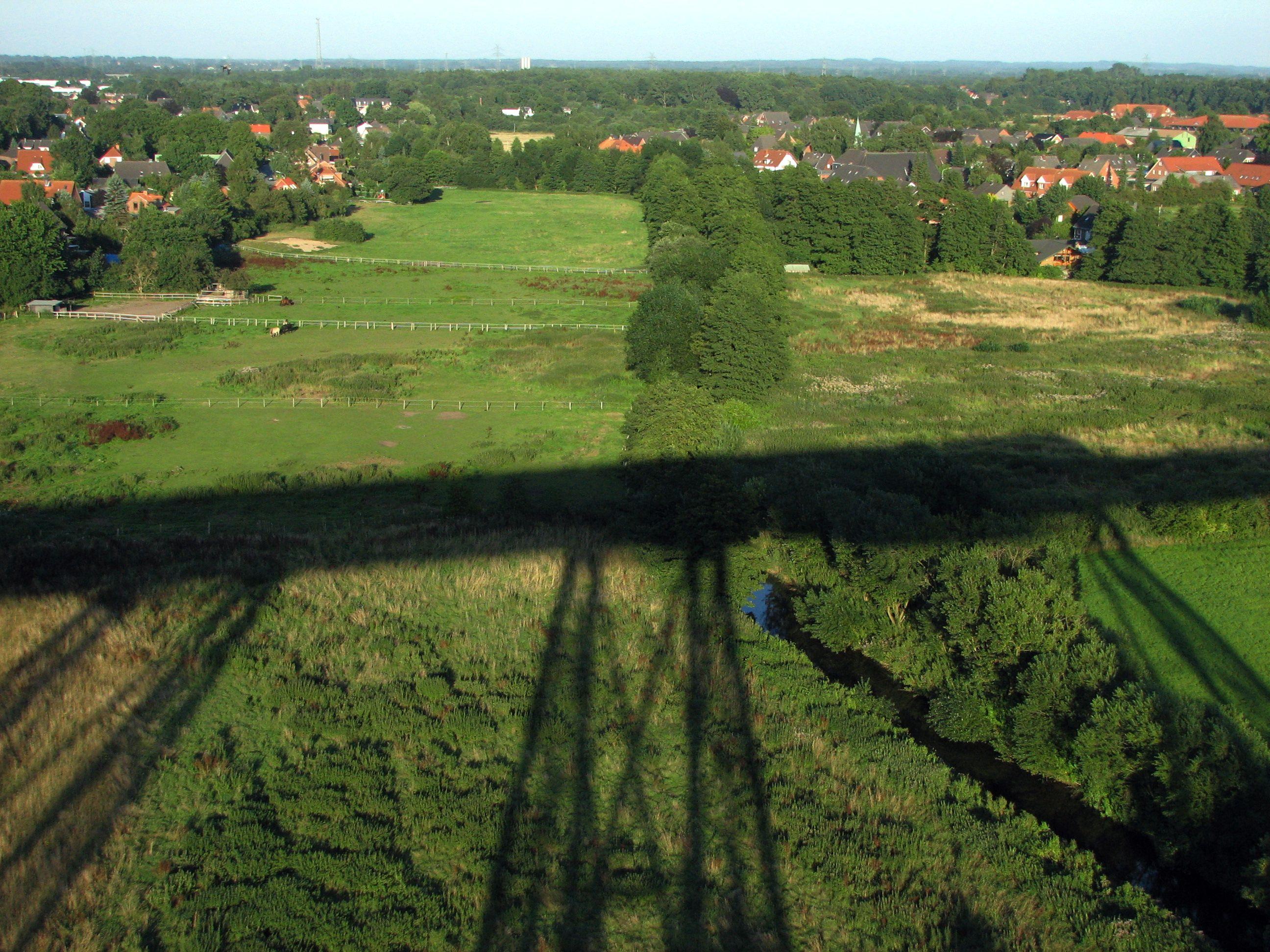 L'ombre d'un haut viaduc métallique projetée sur le paysage verdoyant et relativement plat du Schleswig-Holstein à Rendsburg. Les toitures des habitations voisines sont visibles dans le fond de l'image et se fondent dans la végétation abondante.

The shadow of a high metal viaduct cast over the green and relatively flat landscape of Schleswig-Holstein in Rendsburg. The roofs of neighboring homes are visible in the background of the image and blend into the abundant vegetation.
