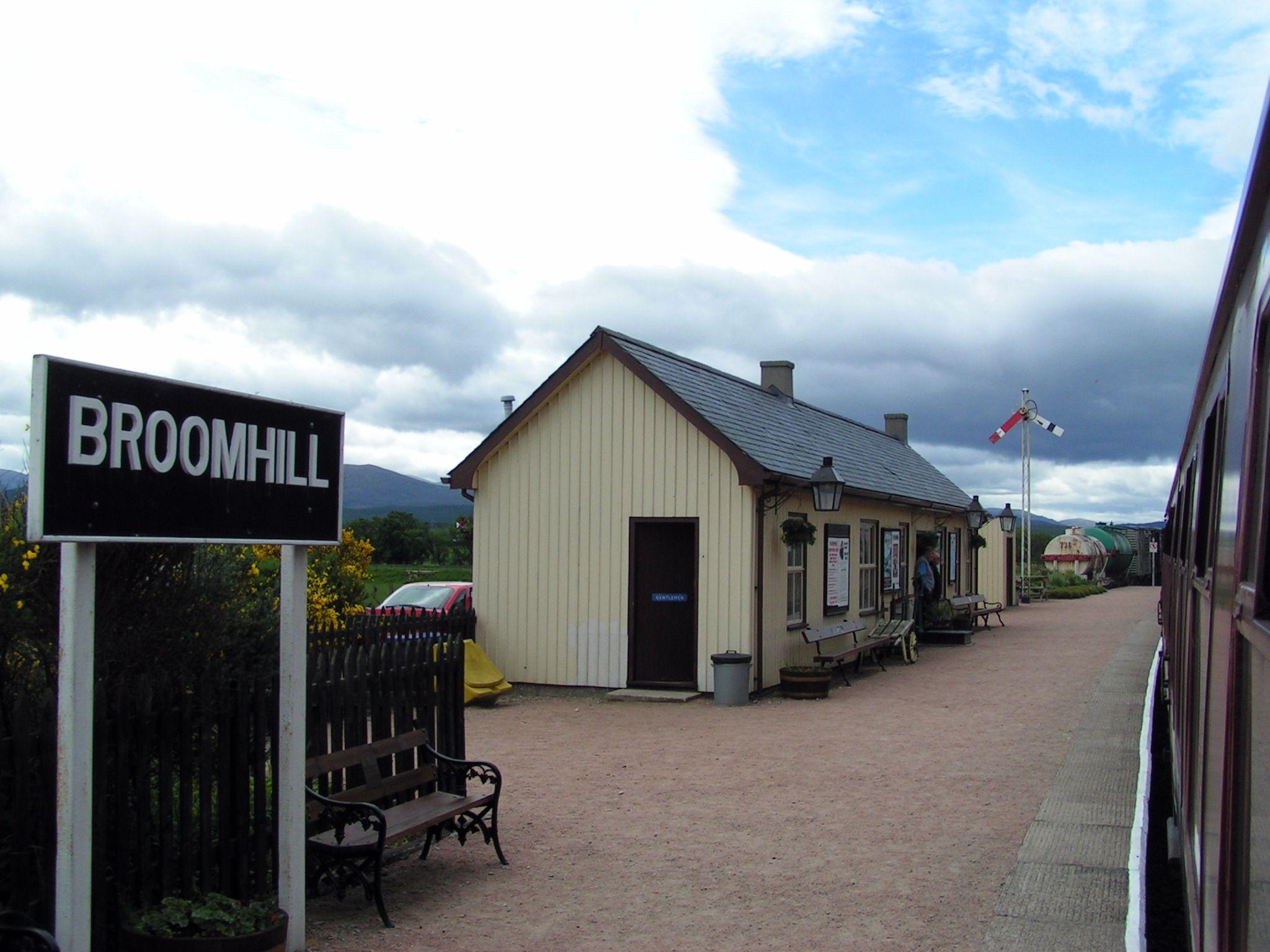 Une petite gare photographiée depuis le train à quai. A gauche le panneau indiquant le nom de la gare Broomhill. Le ciel est nuageux.

A small station photographed from the train at the platform. On the left the sign indicating the name of Broomhill station. The sky is cloudy.