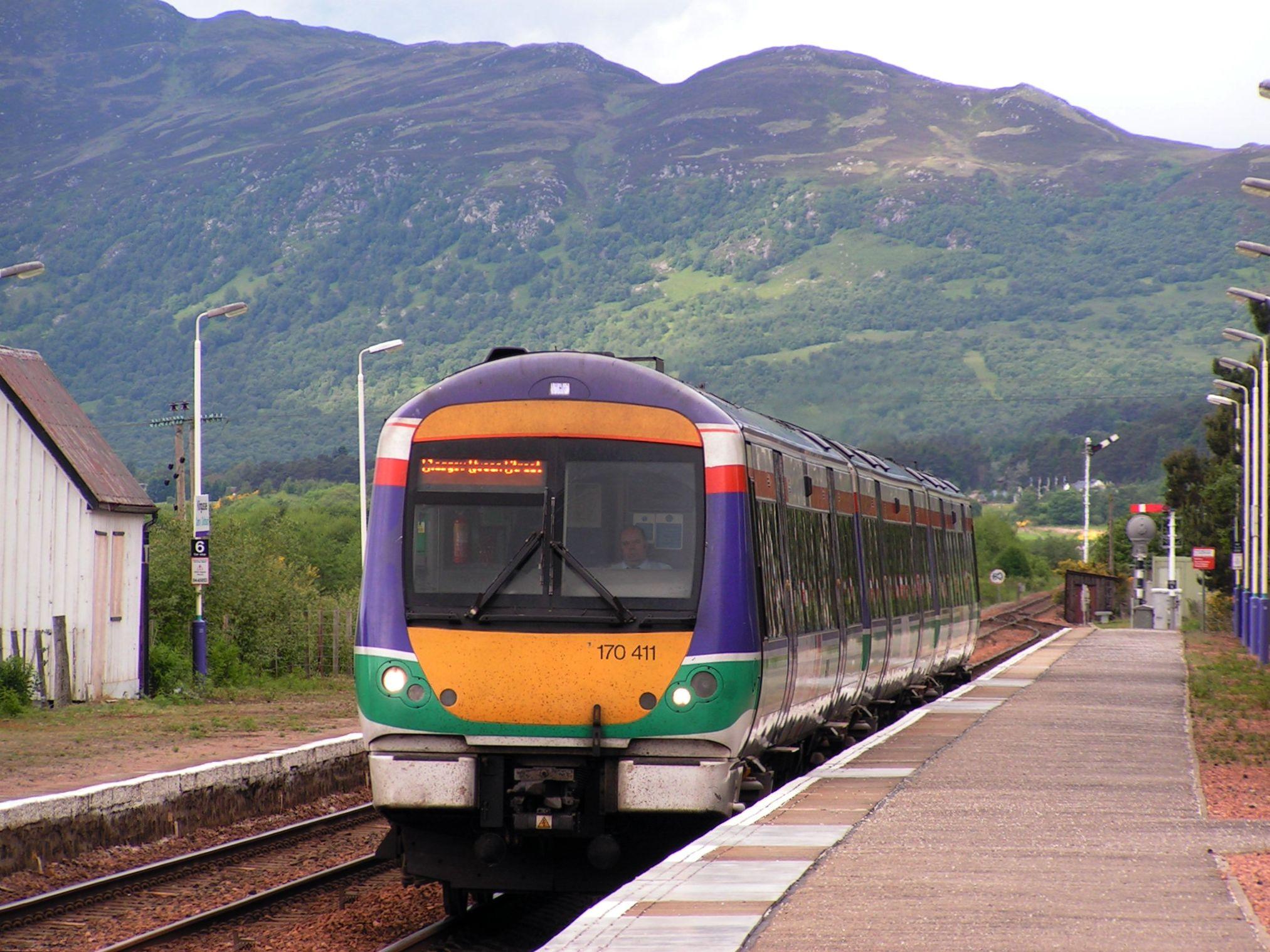 Sur fond des collines typiques des Highlands a Kingussie, un train arrive en gare. Le train a la face avant jaune et porte la livrée faite de bandes rouge, bleu indigo et vert eau.

Against the backdrop of the typical Highland hills in Kingussie, a train arrives at the station. The train has a yellow front and bears the livery made of red, indigo blue and water green stripes.