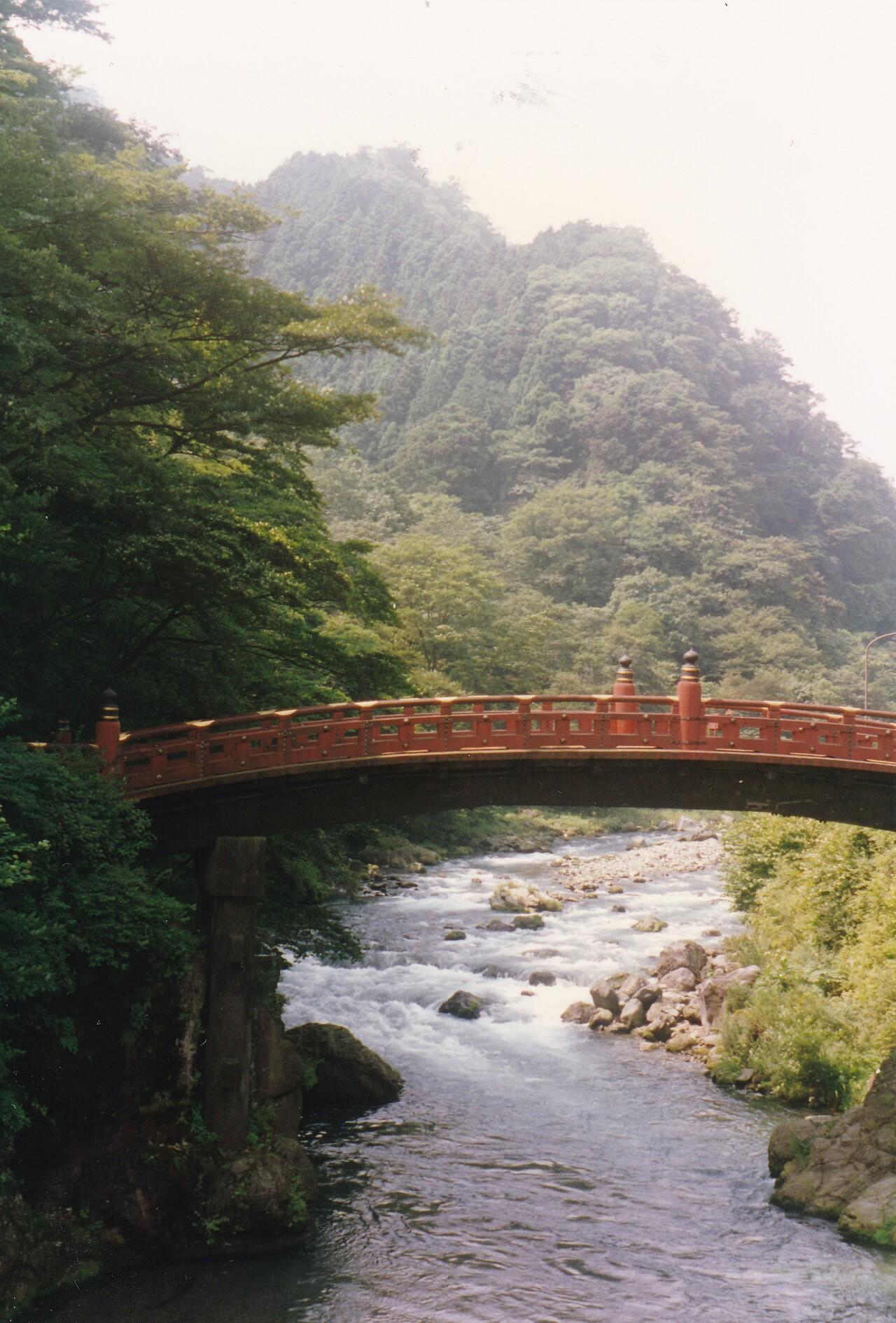 Pont japonais en bois laqué rouge, enjambant la rivière sur fond de collines couvertes d’arbres verts, à Nikko au Japon.