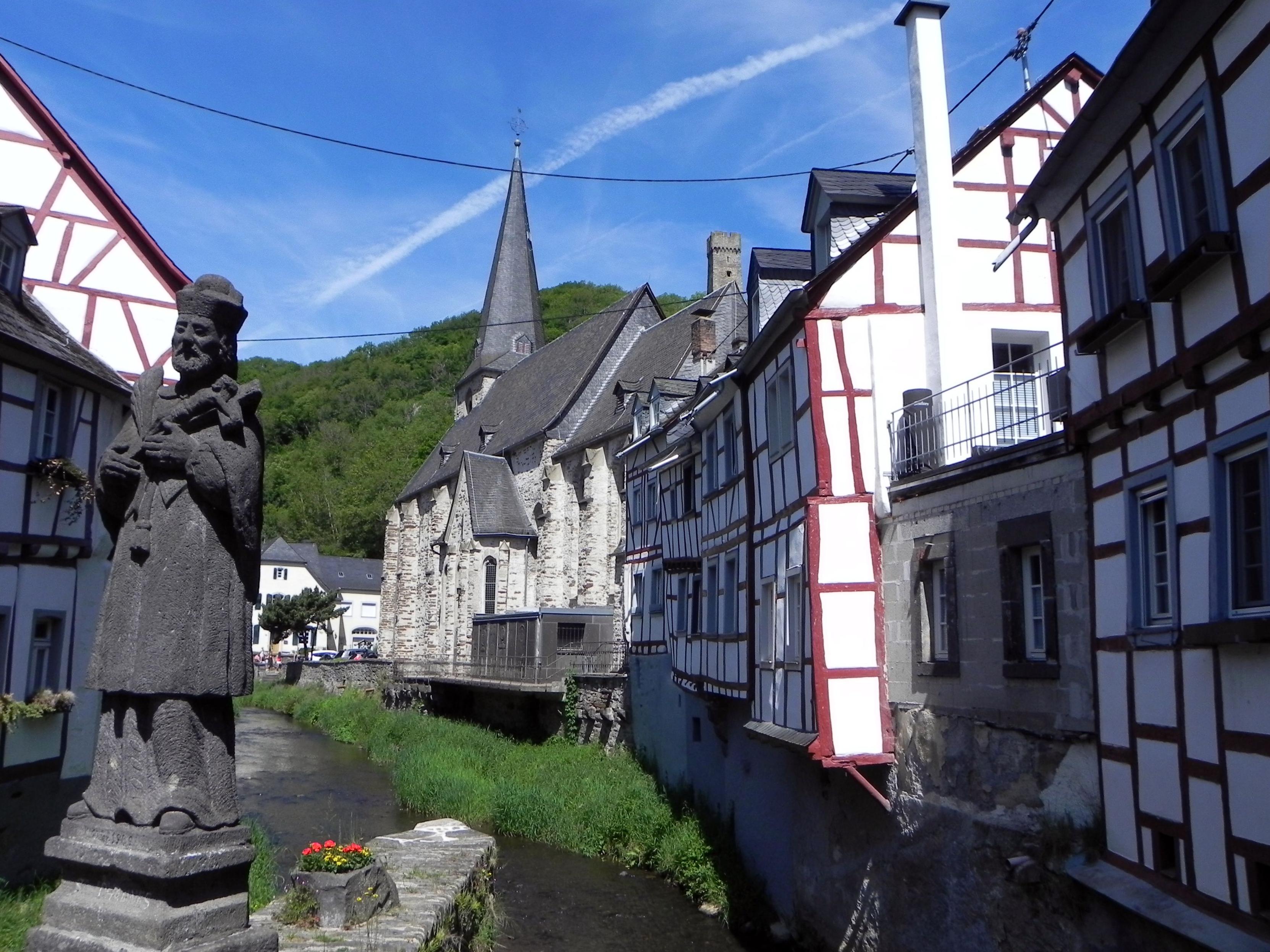 Vue sur une partie des maisons historiques du village de Monreal depuis le petit pont de pierres qui enjambe la rivière locale. Une statue de Jean Nepomuk est placée sur le rebord du pont au milieu de celui-ci. Dans le fond, on distingue l'église du village et tout au fond, l'ancien presbytère qui est un café-restaurant aujourd'hui. A l'arrière, une colline boisée complète le paysage. 

View of some of the historic houses of the village of Monreal from the small stone bridge which spans the local river. A statue of John Nepomuk is placed on the edge of the bridge in the middle of it. In the background, we can see the village church and at the very back, the old presbytery which is a café-restaurant today. At the back, a wooded hill completes the landscape.