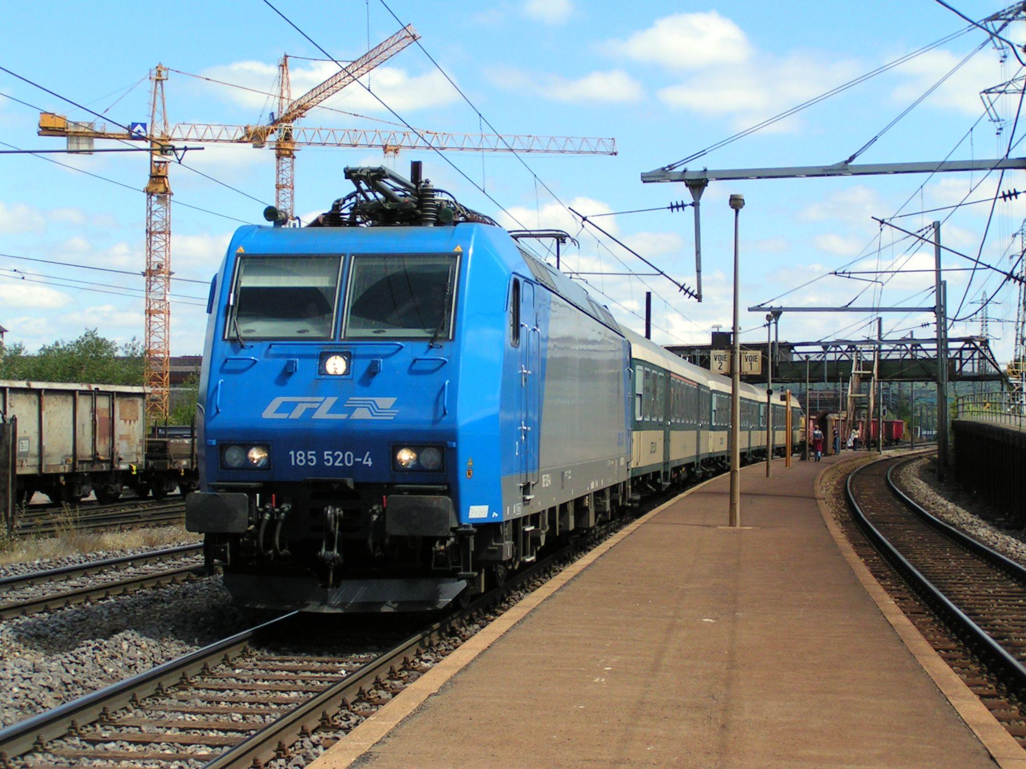 Une locomotive de location série 185 en tête d'une rame de voitures Wegmann à l'arrêt en gare de Belval-Usines. On peut apercevoir la passerelle qui permettait de rejoindre les usines et le quai. 

A locomotive class 185 heading a rake of Wegmann carriages stopping in Belval-Usines. We can see in the background the footbridge giving access to the works and to the platform.