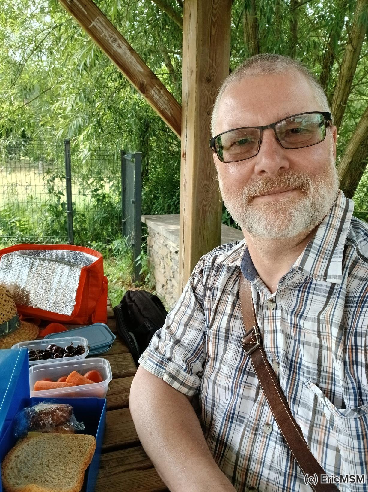 Selfie d’un homme dans la cinquantaine, barbu et portant lunettes et cheveux courts attablé avec une boite à tartines,des tomates, des carottes et des cerises dans deux petites boites. 

Selfie of a 50 something old man with beard, short hair and glasses at a table with his lunch box and two little boxes with vegetables and fruits.