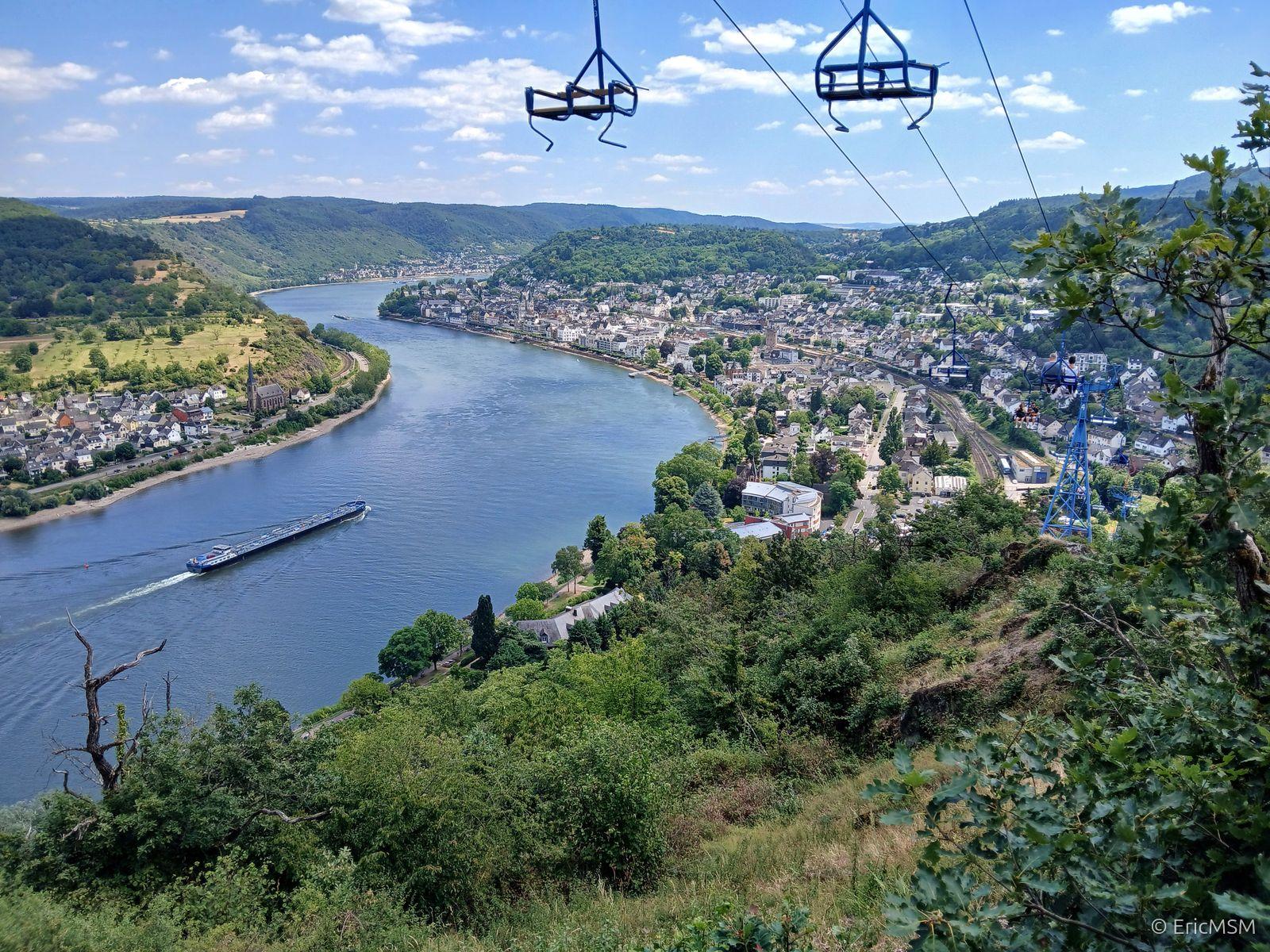 La vallée du Rhin à hauteur de Boppard (à droite) de la photo photographiée depuis le sentier de randonnée Saar-Hunsrück-Steig qui se transforme en sentier approximatif à travers le massif schisteux. Ce sentier fait partie d'un sentier d'escalade. Il suit sur 700 mètres le tracé du télésiège. Une péniche navigue sur le Rhin, un autre village est visible sur l'autre rive à gauche. On peut voir plusieurs sièges du télésièges qui montent et descendent en permanence tout au long de la journée. On peut aussi voir les voies de chemins de fer de la ligne du Rhin (rive gauche). 

The Rhine valley near Boppard (right) in the photo photographed from the Saar-Hunsrück-Steig hiking trail which turns into an approximate trail through the shale massif. This trail is part of a climbing trail. It follows the route of the chairlift for 700 meters. A barge sails on the Rhine, another village is visible on the other bank on the left. Several chairlift seats can be seen constantly moving up and down throughout the day. You can also see the railway tracks of the Rhine line (left bank).