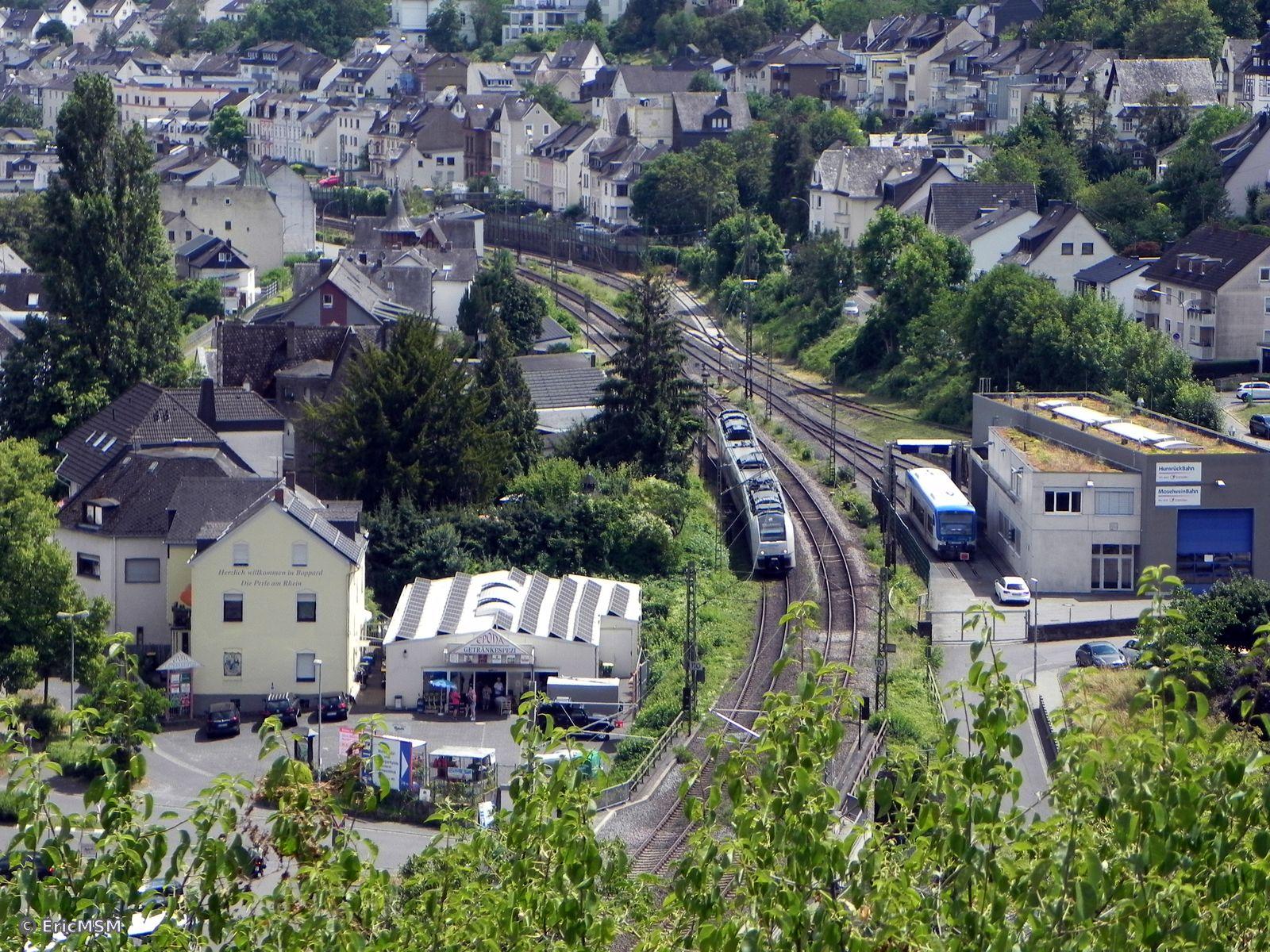 Vue depuis les hauteurs sur les voies de la ligne ferroviaire du Rhin (rive gauche). Un train quitte la gare de Boppard et passe à côté de la remise pour les trains de la ligne vers Emmelshausen. Les voies sont entourées de nombreuses maisons et autres bâtiments.

View from the heights of the tracks of the Rhine railway line (left bank). A train leaves Boppard station and passes the discount for trains on the line to Emmelshausen. The tracks are surrounded by many houses and other buildings.