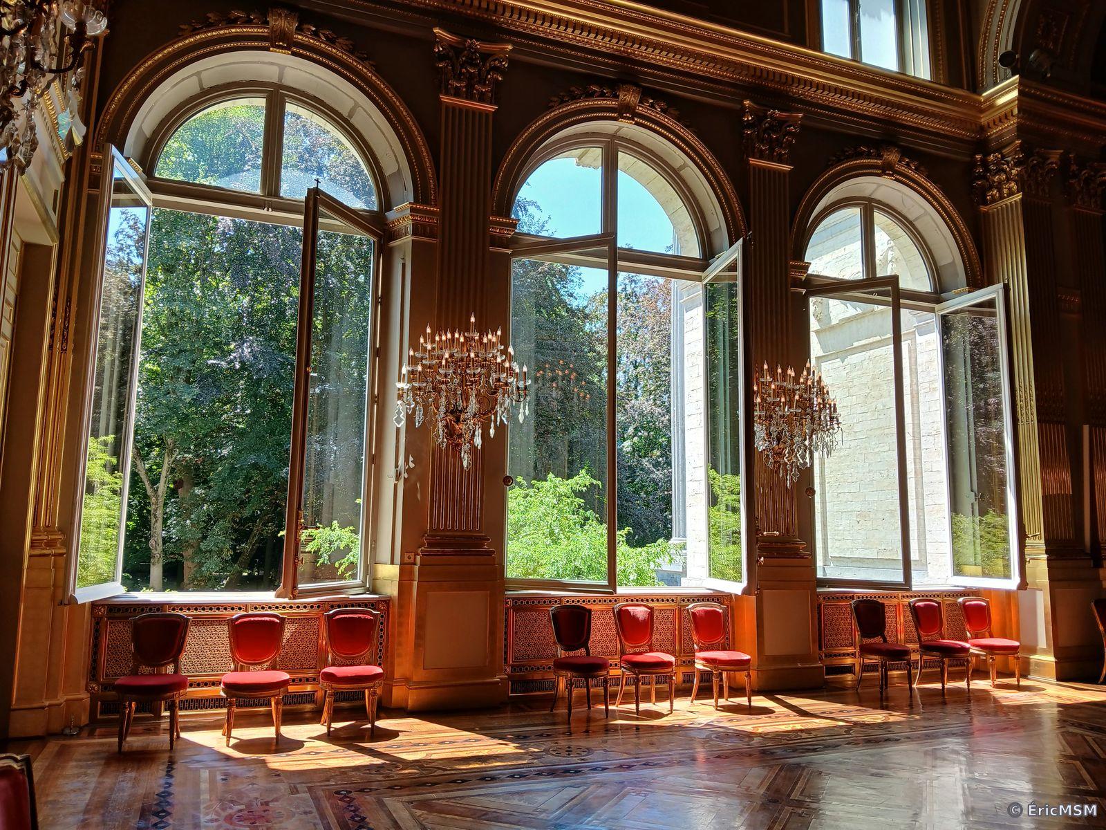 Trois grandes baies vitrées dans une salle du palais royal de Bruxelles, toutes fenêtres ouvertes, il y a beaucoup de dorures, ce qui donne une ambiance très chaude à la photo, elle est prise en contre-jour. On peut voir deux grands lustres et quelques chaises disposées devant les fenêtres. Dans l'arrière-plan, on distingue des arbres.

Three large bay windows in a room of the royal palace of Brussels, all windows open, there is a lot of gilding, which gives a very warm atmosphere to the photo, it is taken against the light. We can see two large chandeliers and a few chairs arranged in front of the windows. In the background, we can see trees.