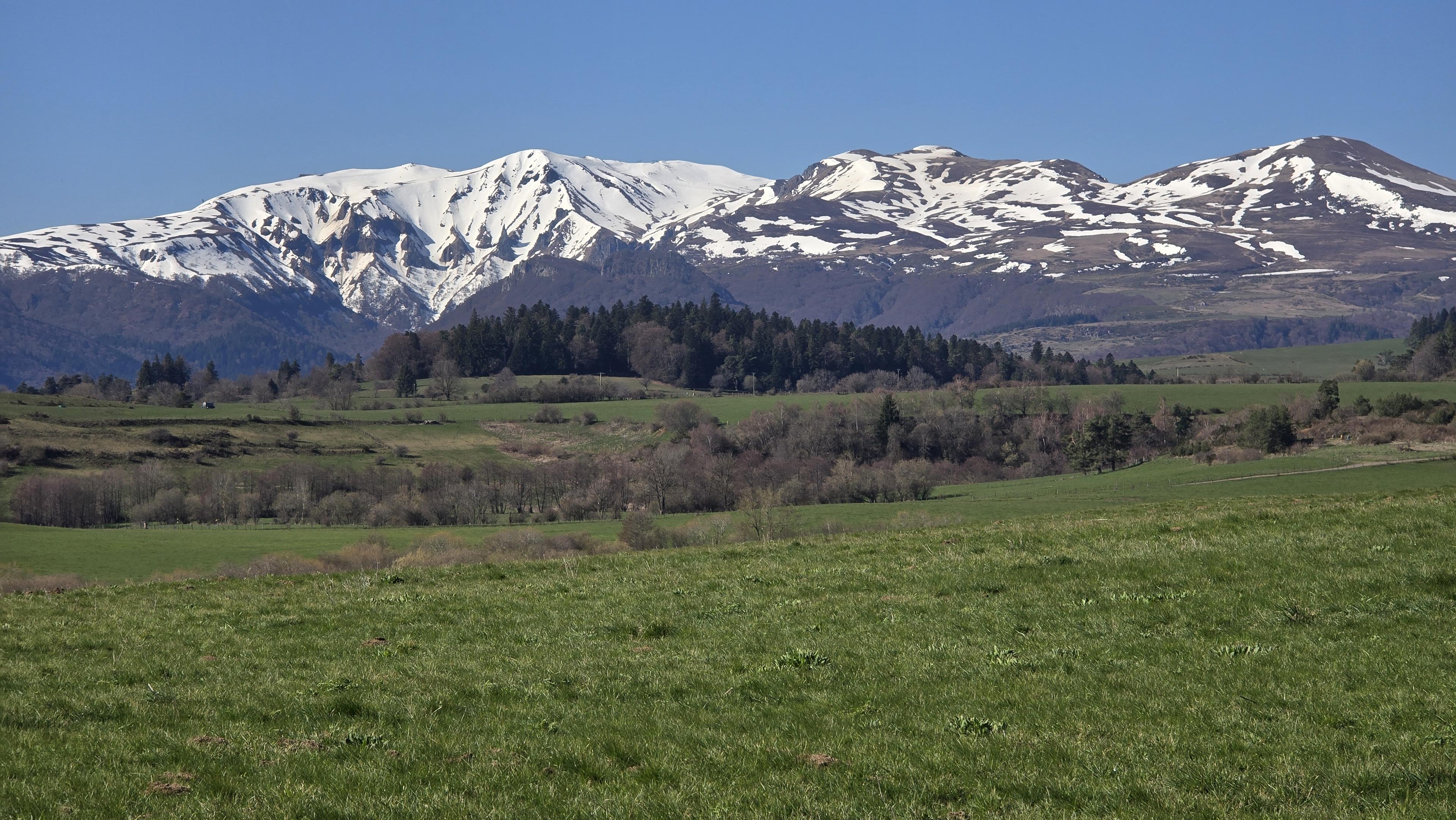 Vue panoramique sur le massif du Sancy en Auvergne. Les sommets sont enneigés, une prairie verte est au premier plan.