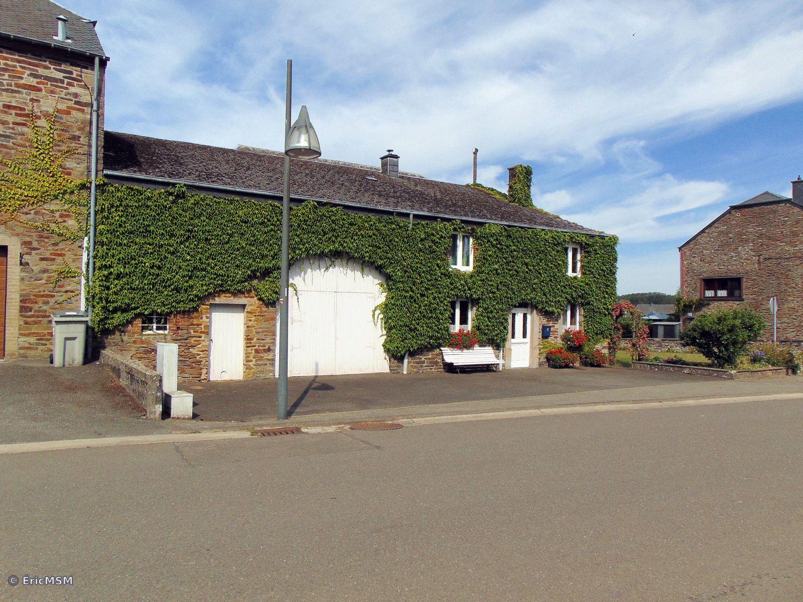 Une jolie maison à la façade bien végétalisée sur une grande partie, une grande porte blanche est située au milieu du bâtiment et un banc, blanc lui aussi, se trouve sous une fenêtre.

A pretty house with a well-vegetated facade over a large part, a large white door is located in the middle of the building and a bench, also white, is under a window.