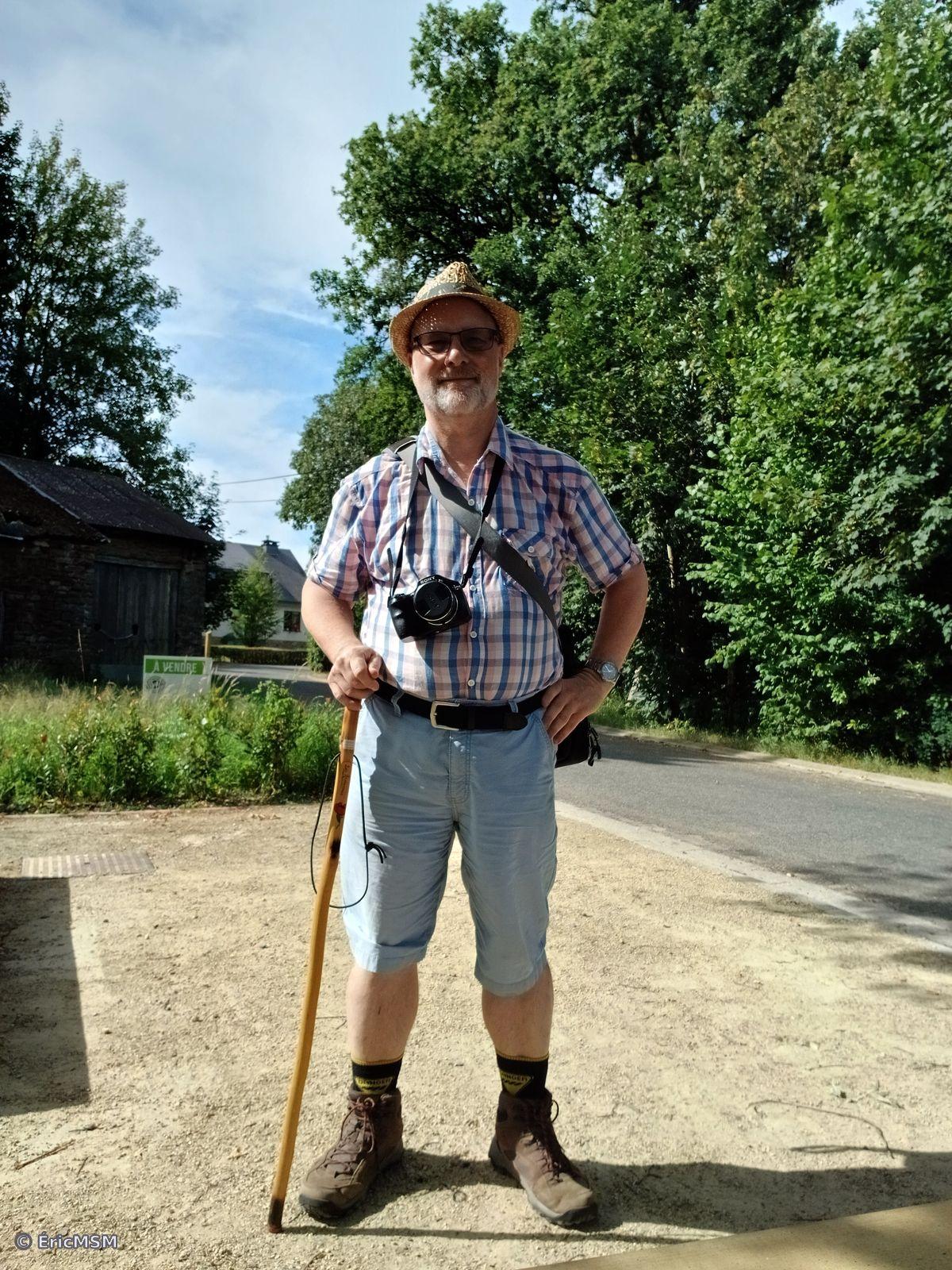 Le randonneur satisfait! Selfie d'un homme dans la cinquantaine portant un chapeau de paille, un appareil photo autour du cou, un baton de marche dans la main droite (à gauche sur la photo), il porte un bermuda bleu pâle et des bottines de marche. L'arrière-plan est composé essentiellement d'arbres. 

The satisfied hiker! Selfie of a man in his 50s wearing a straw hat, camera around his neck, walking stick in his right hand (left in photo), he wears pale blue Bermuda shorts and walking boots. The background is mostly trees.