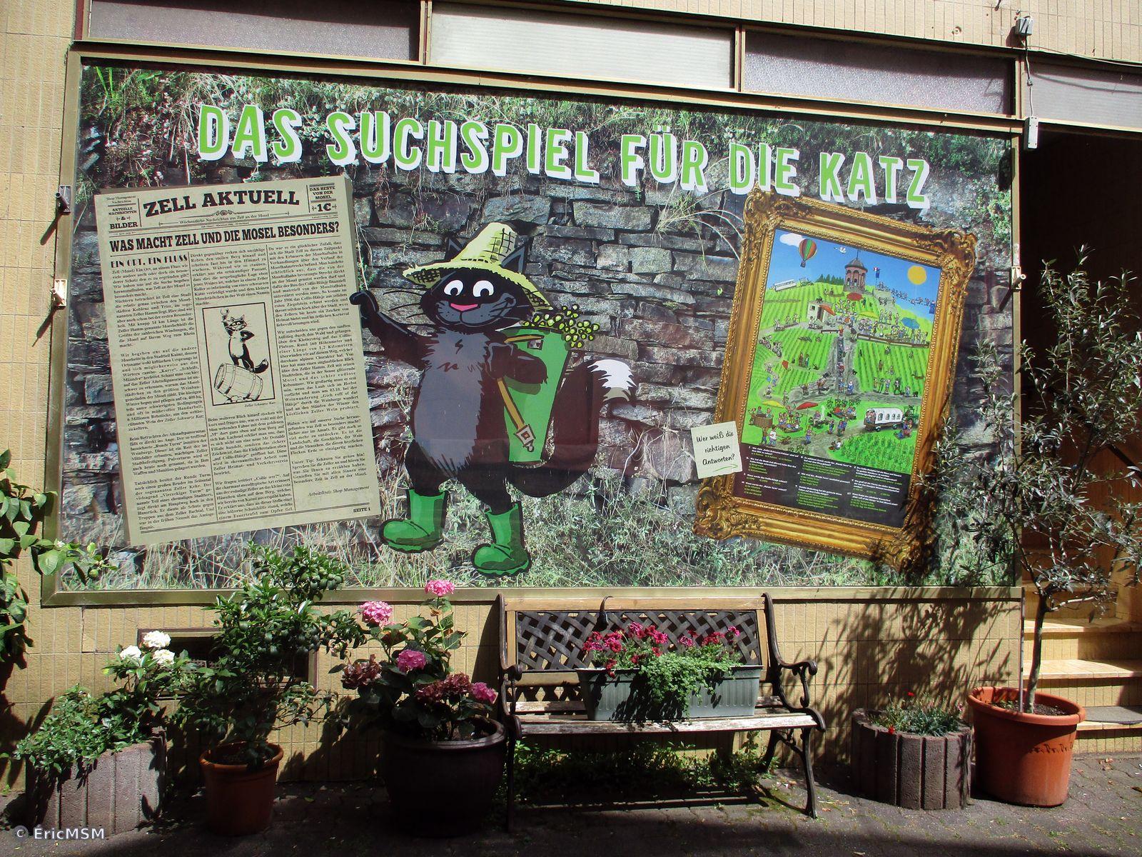 Un panneau explicatif à propos d'un jeu avec le célèbre chat noir de Zell (Mosel) Il tient un journal à gauche et à droite, on voit une tour célèbre représentée dans un tableau. Il y a un banc et des pots de fleurs sous le panneau.

An explanatory panel about a game with Zell's famous black cat (Mosel) He keeps a diary left and right, we see a famous tower represented in a painting. There is a bench and flower pots under the panel.