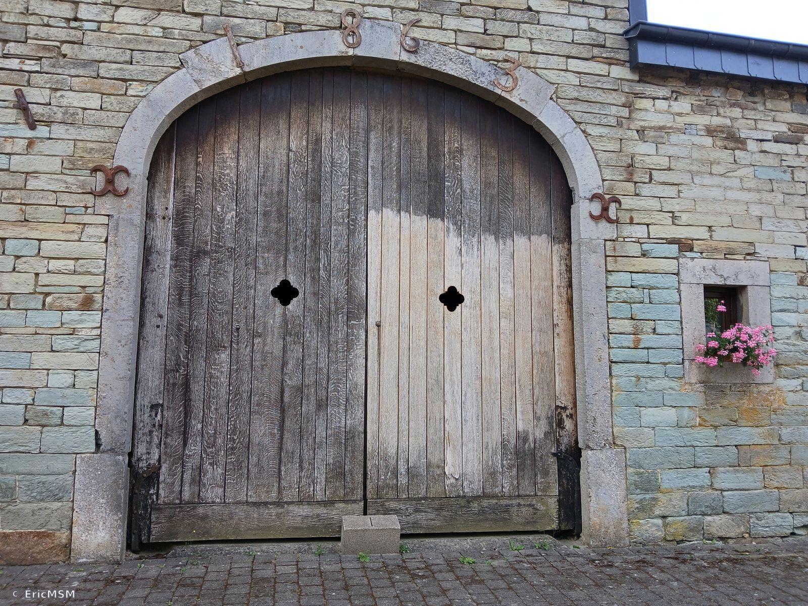 Une grand porte en bois à double battant avec des losanges découpés à mi-hauteur, des chiffres indiquent 1863 au-dessus du porche. Le bâtiment est en pierre d'Ardenne.

A large double-leaf wooden door with diamonds cut halfway up, figures indicate 1863 above the porch. The building is made of Ardennes stone.