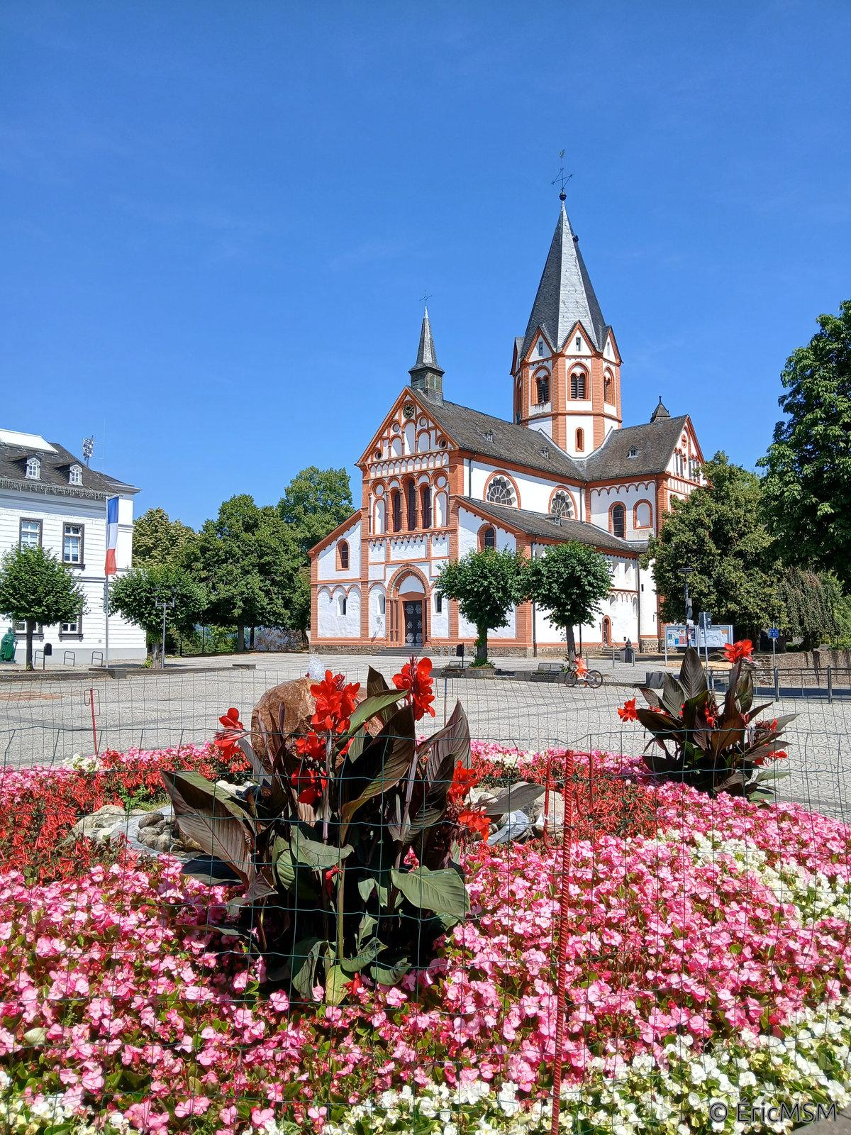 Une place dans une petite ville sous le soleil et un ciel bleu sans nuage, un beau massif fleuri est visible en premier plan, à l'arrière-plan, une église avec un cloché octogonal peinte en blanc dont les arêtes sont rehaussées par la couleur terre de Sienne. A gauche, un bout de l'hôtel de ville est visible.

A square in a small town under the sun and a cloudless blue sky, a beautiful flower bed is visible in the foreground, in the background, a church with an octagonal bell tower painted white whose edges are enhanced by the color land of Siena. On the left, part of the town hall is visible.