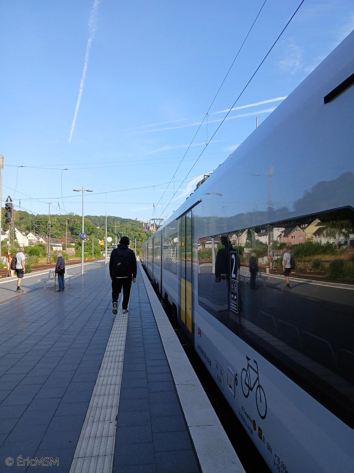 Un train en gare avec dans le fond une imposante église sur une colline boisée. Une personne se déplace le long du train et deux autres attendent de l'autre côté du quai.

A train at the station with an imposing church in the background on a wooded hill. One person is moving along the train and two others are waiting on the other side of the platform.