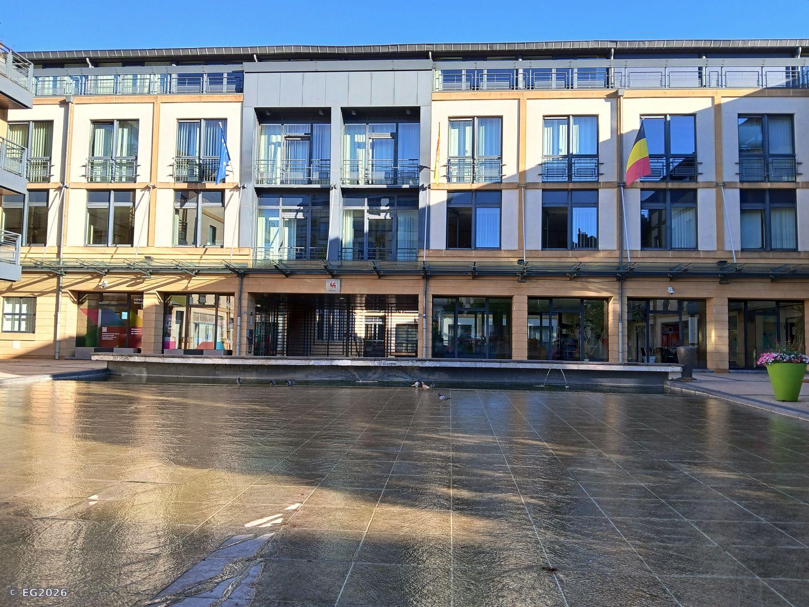 Un bâtiment moderne avec beaucoup de fenêtres et un plan d'eau à l'avant-plan qui fait la joie des pigeons. 

A modern building with lots of windows and a body of water in the foreground that delights pigeons.