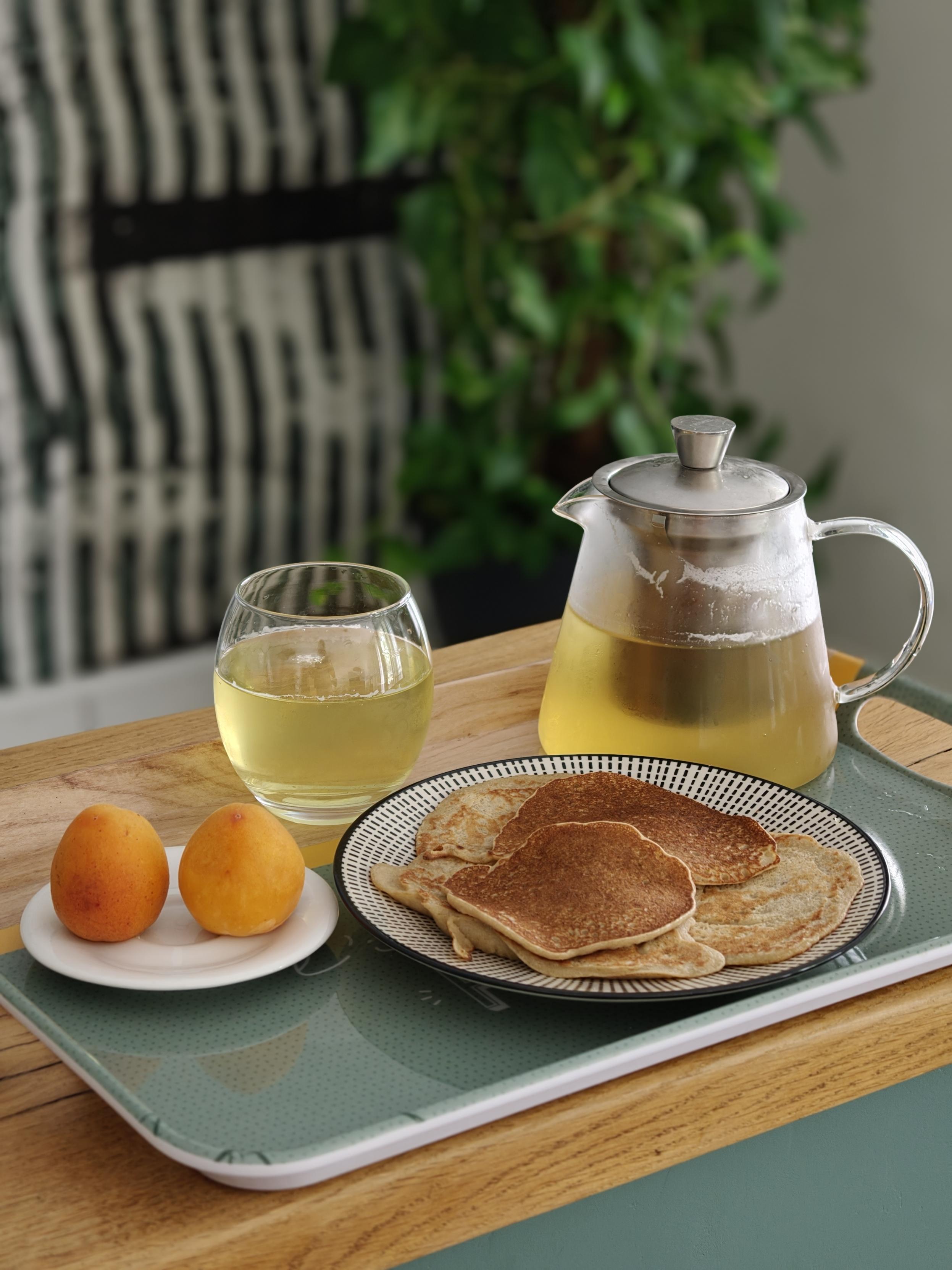 Plateau avec une assiette de pancakes banane, cannelle, rhum, deux abricots et une théière de thé vert orange douce.